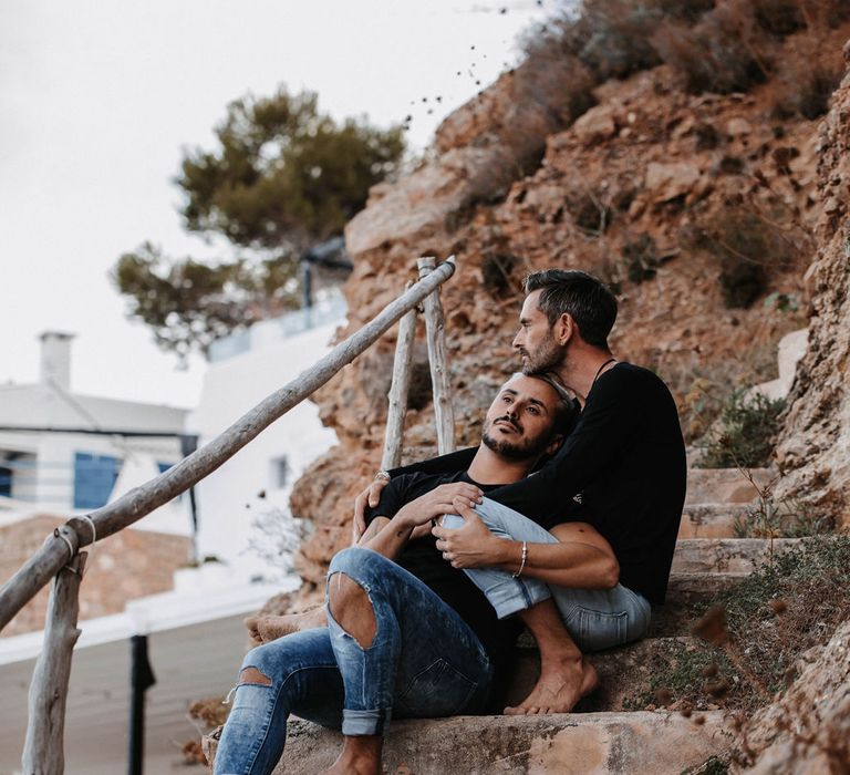 Grooms sit on rocks together in Ibiza