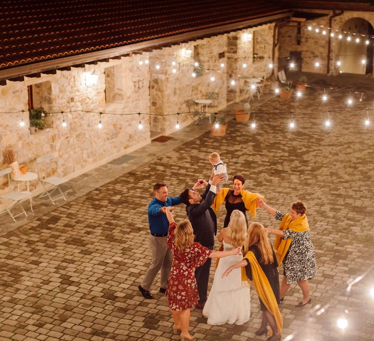 Guests dancing in a circle around the bride and groom in a courtyard, as the groom lifts his baby son into the air