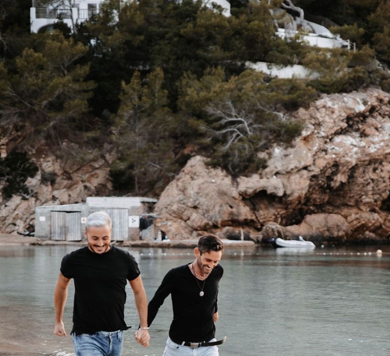 A gay couple walk along the beach having fun for  a pre wedding photo shoot. They both wear black tops and have jeans rolled up at the ankles. Photography by Stephanie Shenton. 