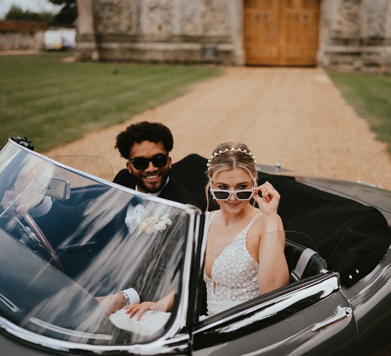 Bride in white sunglasses with a pearl headband sitting in a classic wedding car with her husband in a tuxedo 