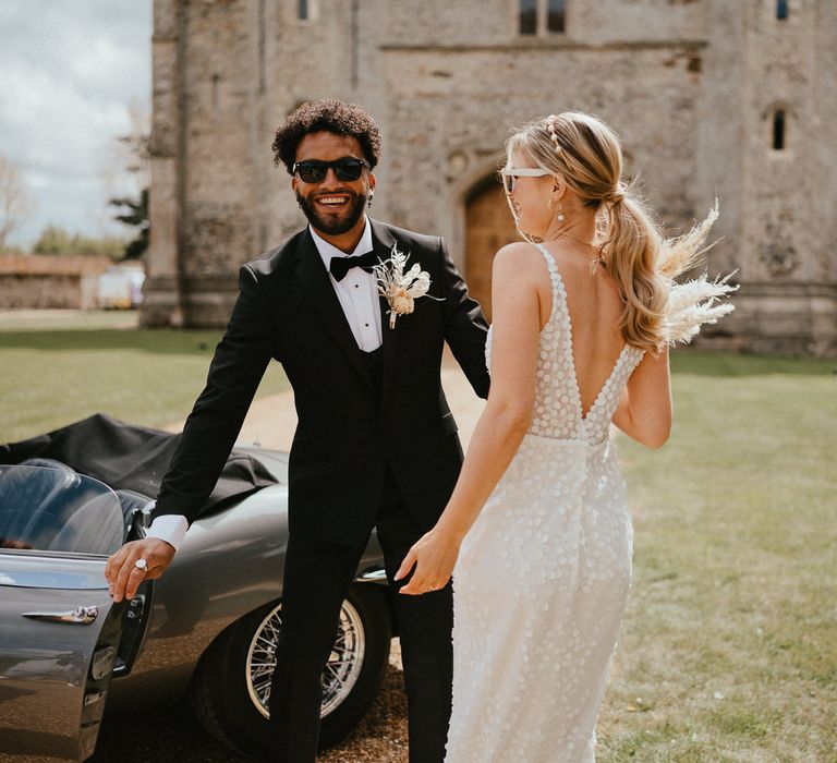 Groom in a tuxedo with bow tie, sunglasses and dried flower buttonhole holding the car door open for his bride in an appliqué wedding dress