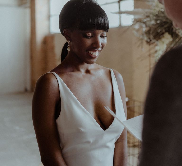 A Black bride smiles as the groom reads his vows. She wears a low cut sleek white dress and has her hair tied back in a low ponytail. She has a fringe and wears a nose ring. 