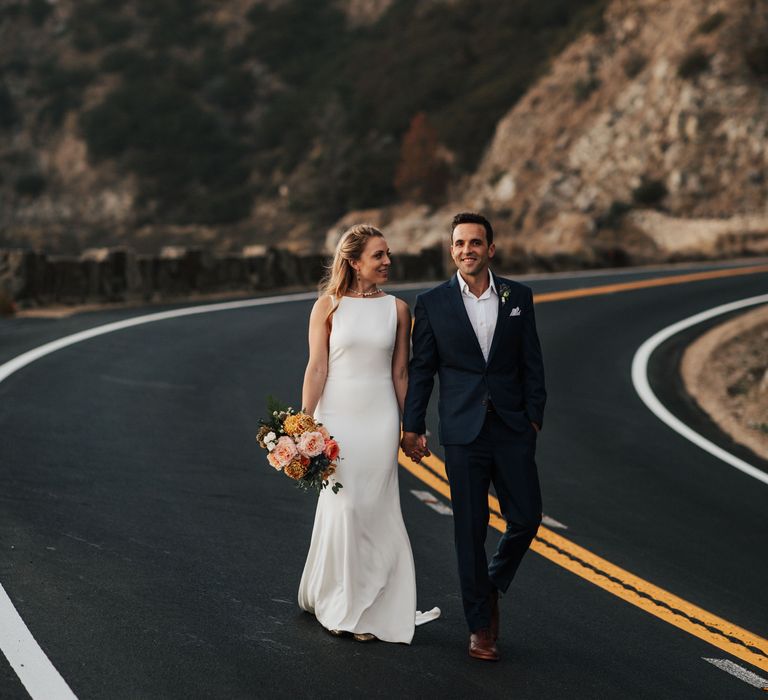 Bride & groom walk along the road with one another as bride carries peach bouquet