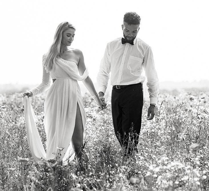 Black and white portrait of the bride and groom holding hands in a field. The bride wears a wrap wedding dress with front split and one shoulder detail 