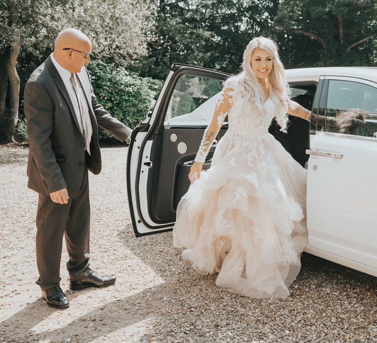 A bride in a tiered tulle wedding dress steps out of a white wedding car. 