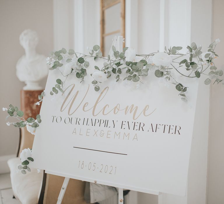 White wedding welcome sign on easel reading 'Welcome To Our Happily Ever After Alex & Emma 18.05.2021' with green foliage decoration