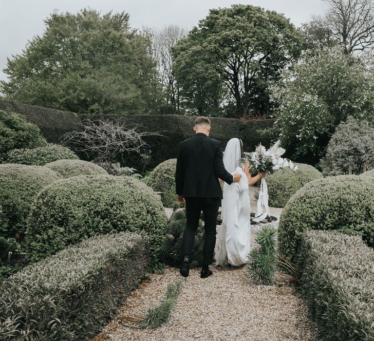 Groom in black tuxedo holds the train of bride's white Pronovias wedding gown as they walk through the gardens at Came House Dorset 