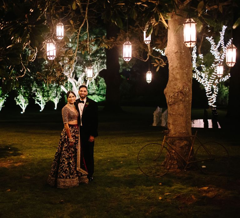 Bride & groom stand outdoors whilst lights twinkle around them during the night