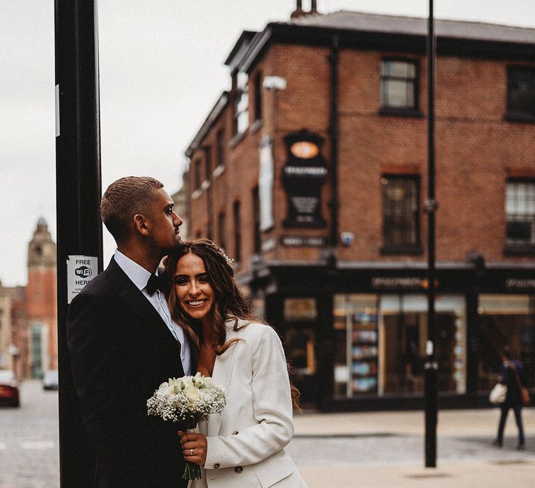 Bride in a wide leg trouser suit holding a white flower bouquet embracing her groom in a black tuxedo 