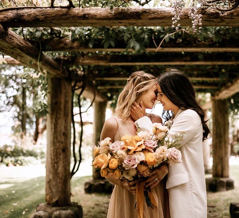 Bride in a white suit kissing her bride in a blush pink wedding dress under a pergola 