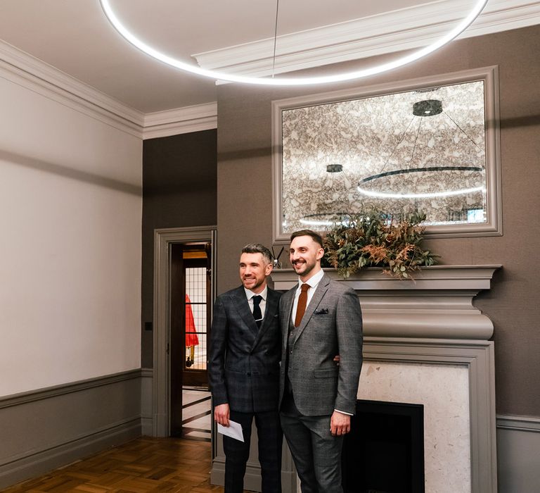 Grooms stand within Old Marylebone Town Hall below neon light for their wedding ceremony