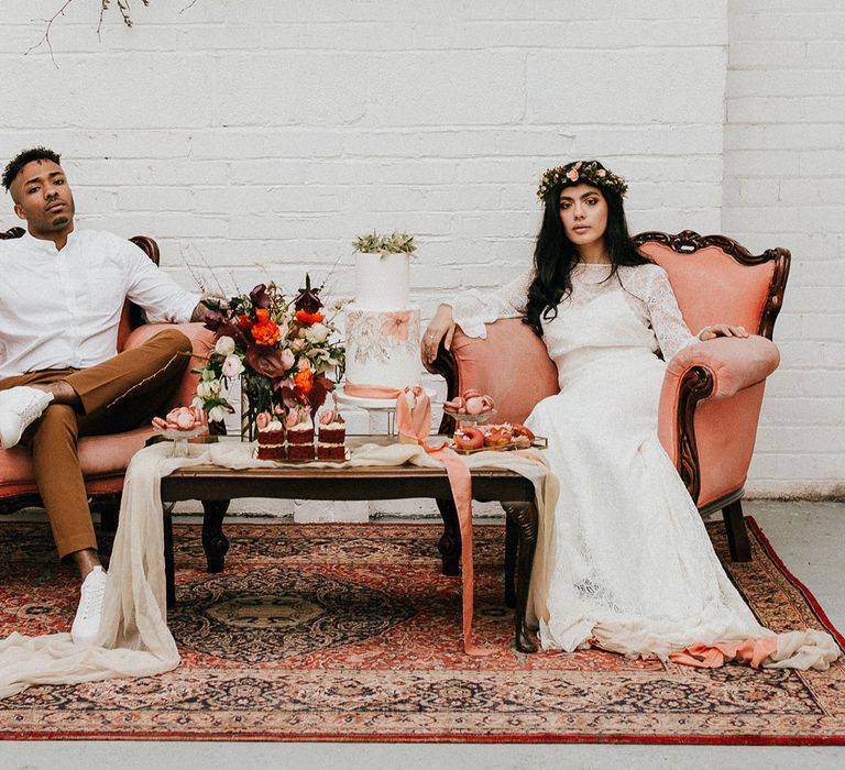 Bride in a lace wedding dress and flower crown with her groom in orange trousers and white shirt sitting on a pink chairs with a dessert table in front of them