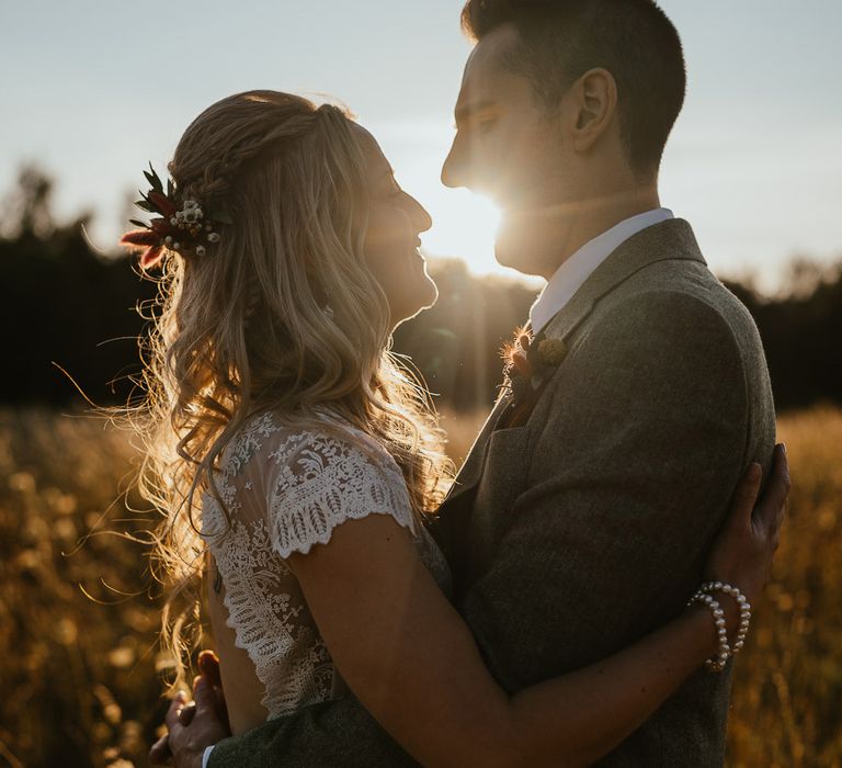 Bride in lace top open back wedding dress with blond curls and flowers in her hair hugs groom in grey herringbone suit in field at golden hour during late summer wedding at Wellington Wood Norfolk