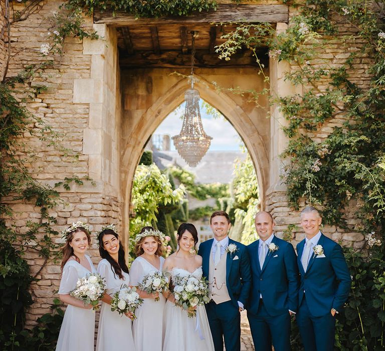 Bride & groom stand with their wedding party in front of archway complete with hanging chandelier 