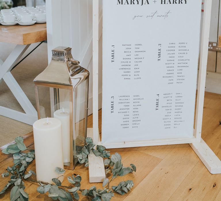 White fabric wedding table plan on white wooden stand with large candles and foliage for summer wedding at Primrose Hill Farm wedding