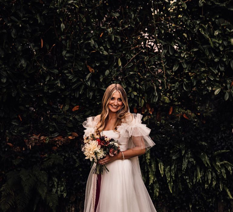 Bride in white Halfpenny London Mayfair dress stands outside holding mixed bridal bouquet with burgundy ribbon before church wedding ceremony in Cornwall