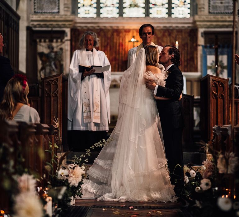 Bride in white Halfpenny London Mayfair dress and veil kisses groom in three piece suit at the altar during church ceremony in Cornwall 