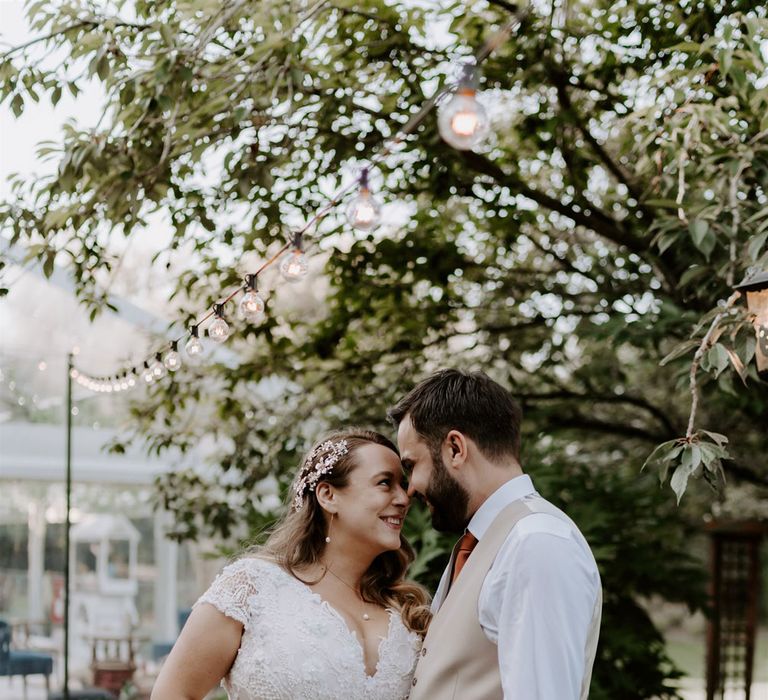 Bride in homemade lace wedding dress with pearl floral hair pin smiles at groom in beige suits trousers and waistcoat as they stand in garden after wedding ceremony