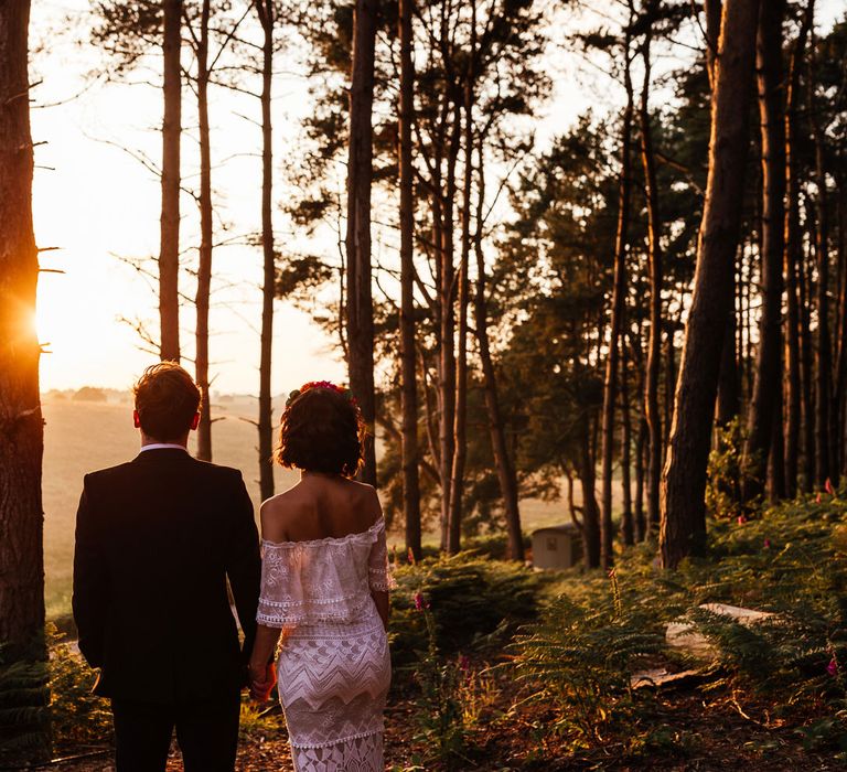 Bride in off the shoulder Grace Loves Lace wedding dress and flower crown stands looking towards the sun holding hands with groom in black suit during golden hour