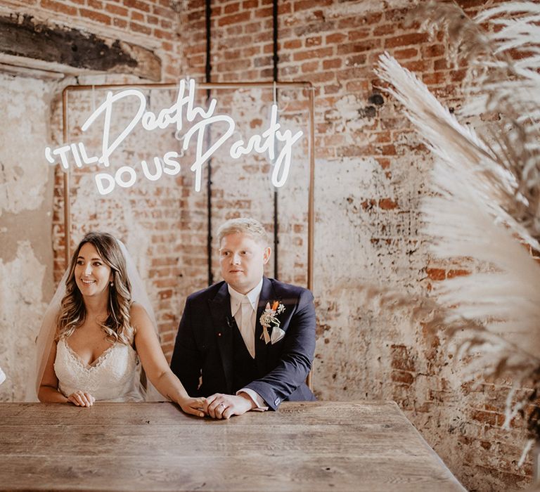 Bride & groom sit under neon sign at wooden table in barn complete with brick wall behind them