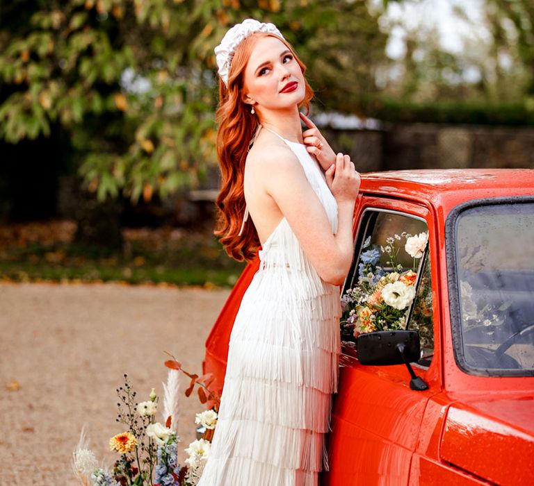 Bride with long curled hair wearing chunky white bridal headband and long fringed wedding dress lean son red car filled with colourful florals