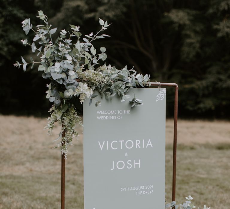 Welcome sign complete in sage green held by brass stand and surrounded by foliage