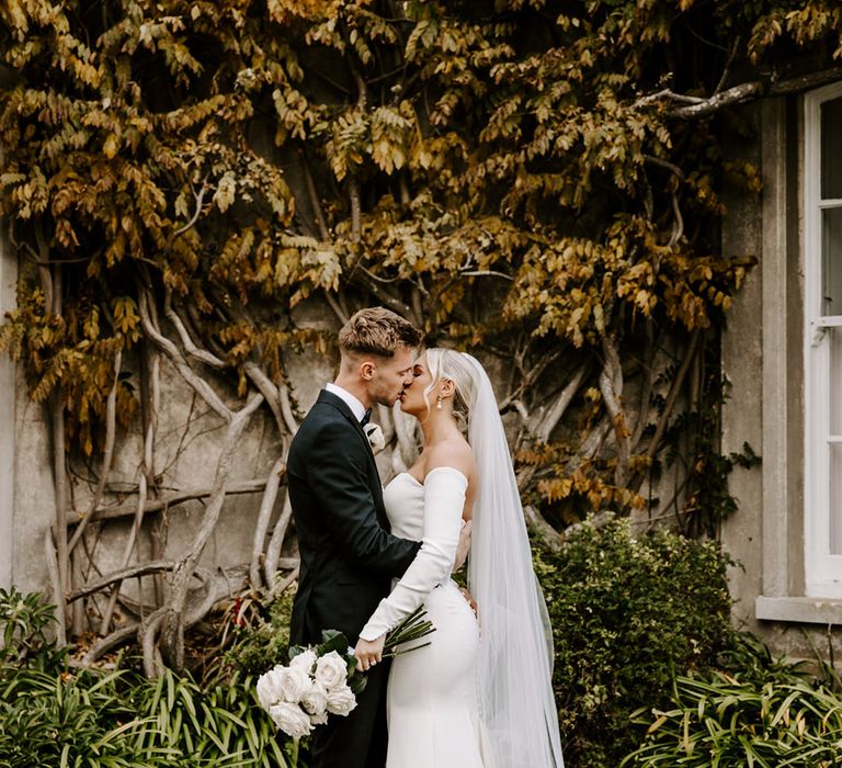 Groom in a tuxedo kissing his bride in a fitted wedding dress with a long cathedral length veil and white rose bouquet 