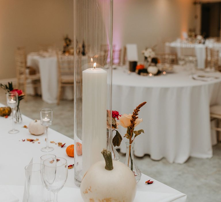 Pillar candle in tall glass holder complete with white pumpkin on white tablecloth 