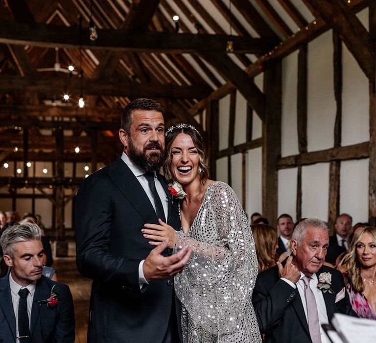 Bride in a silver sequin wedding dress laughing during the barn wedding ceremony with her groom in a navy suit 