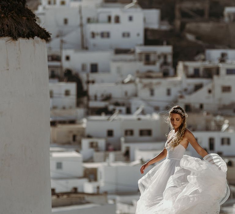 Bride stands on rock as she holds her Watters wedding gown as Greece can be seen in the background