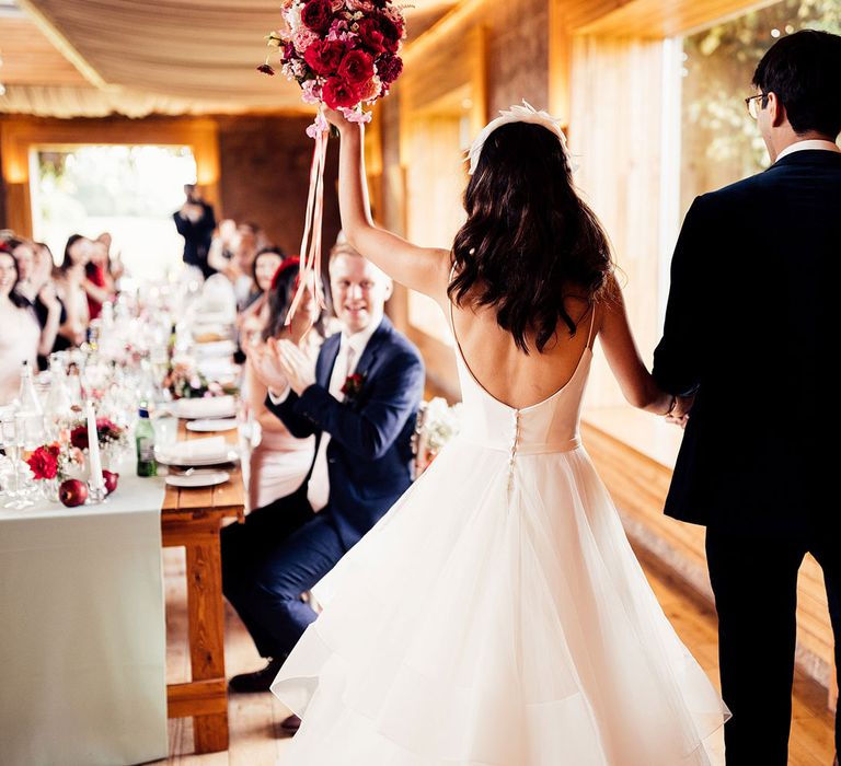 Bride in low back wedding dress with burrow detains and tulle skirt walks into wedding reception with groom in dark suit as she holds up pink and red bouquet