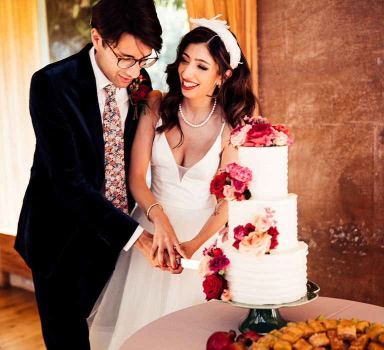 Laughing bride in wedding dress with plunging neckline and feathered headband cuts three tier white wedding cake with groom in dark suit and floral buttonhole