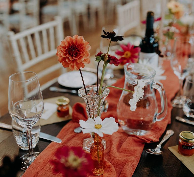 Orange table runner lines wooden table alongside brightly coloured flowers 