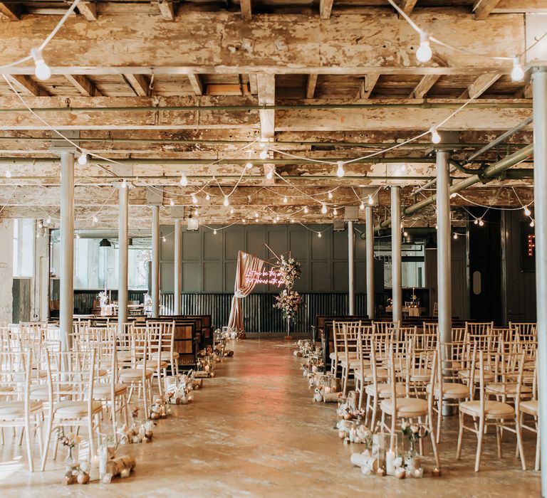 Industrial wedding ceremony room with pillars, wooden chairs, pastel florals and pink neon wedding sign