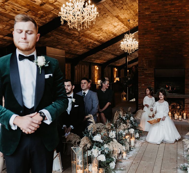 Flower girls in lace white tulle princess dresses walk down the aisle whilst the groom nervously waits