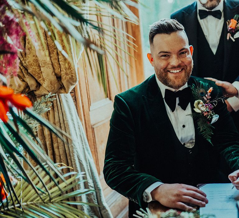 Groom in green velvet suit jacket and black bow tie with large peacock feather boutonnière signs the register with white feather pen