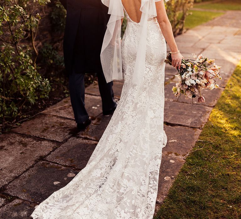 Bride in lace scoop back wedding dress with train holding hands with groom in morning suit during golden hour