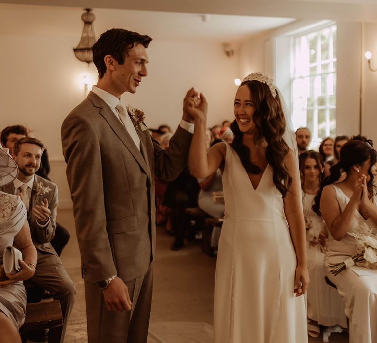 Bride and groom smile at each other as they raise their arms after getting married at Aswarby Rectory