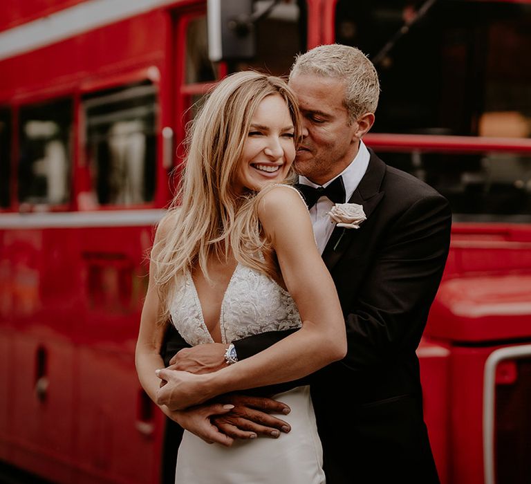 Groom in black tie hugs the bride from behind as they stand in front of their red double decker wedding bus transport 