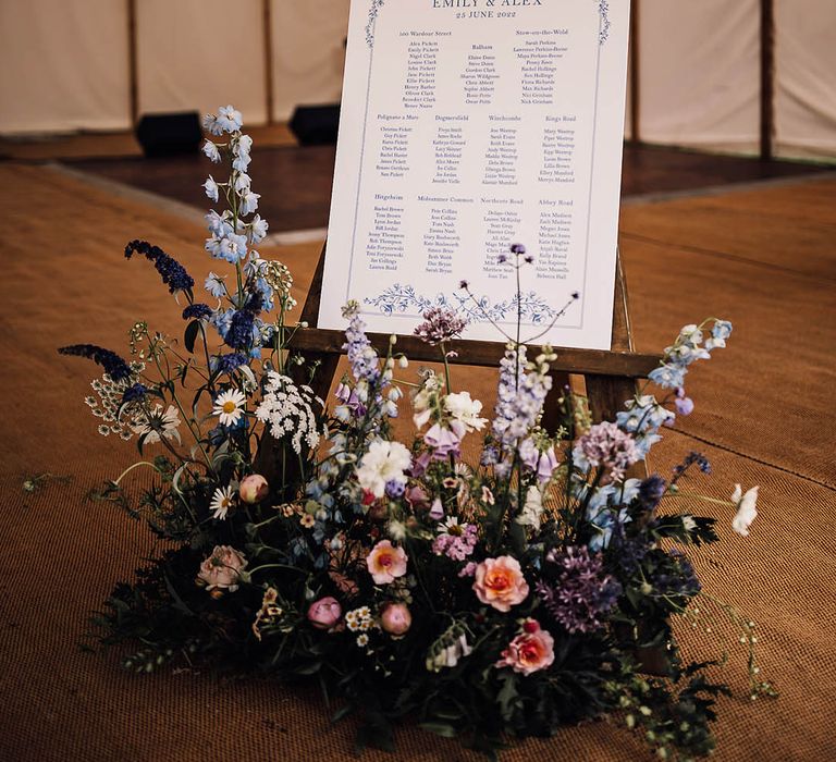 Blue and white wedding stationery table plan sign on an easel with blue and purple wildflowers laying in front 