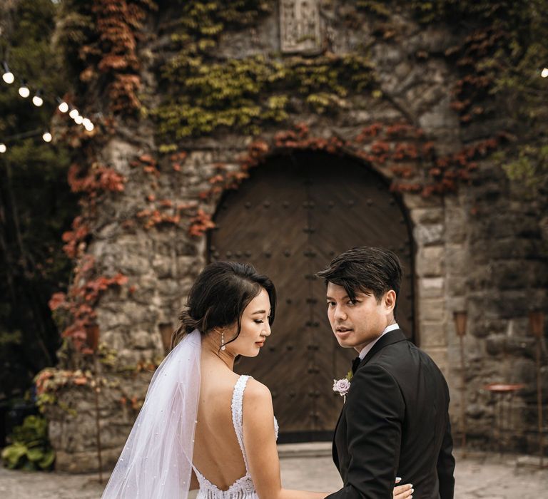 Bride wears floor-length veil with pearl embellishment and stands beside her groom who wears black tie 