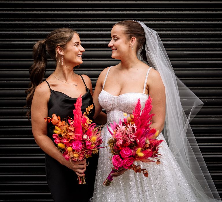 Bride wearing sparkly wedding dress carries brightly coloured dried floral bouquet filled with roses and pink pampas grass stood beside her bridesmaid in a silk black bridesmaid dress 