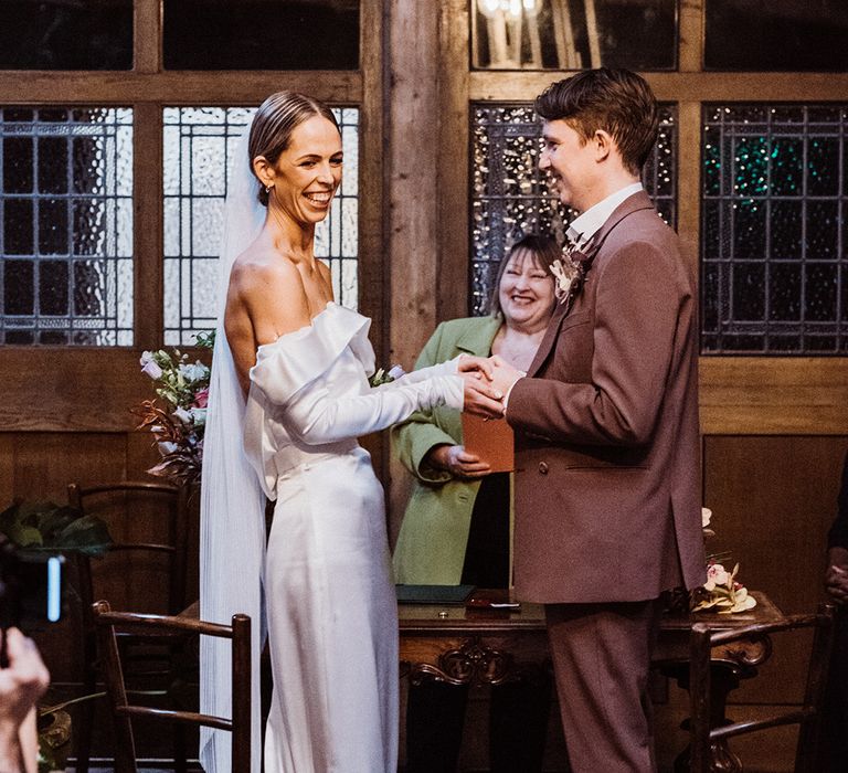 Bride and groom smile widely and hold hands as they participate in their wedding ceremony at Clapton Country Club 