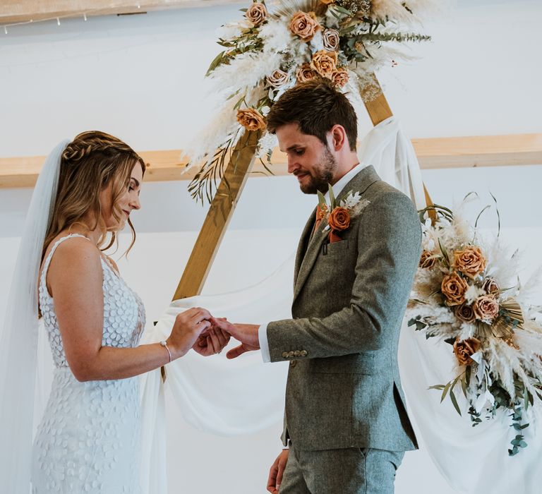 Bride in sleeveless Made With Love Bridal dress with white embellishments and groom in grey suit with orange tie standing by wooden triangle arch with dried flower arrangements