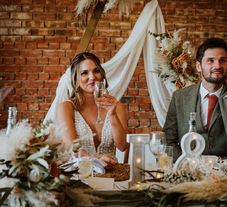 Bride in sleeveless Made With Love Bridal dress with white embellishments and plunge neckline and groom in grey suit with burnt orange tie, pocket square and boutonniere raising their glasses at The Green Cornwall 