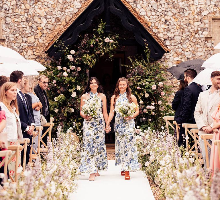 Bridesmaids holding hands in white and blue floral dresses walk down the aisle together 