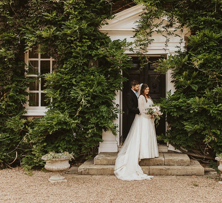 Groom in a morning suit with a pale blue tie embraces the bride from behind as they pose in front of the country house venue 