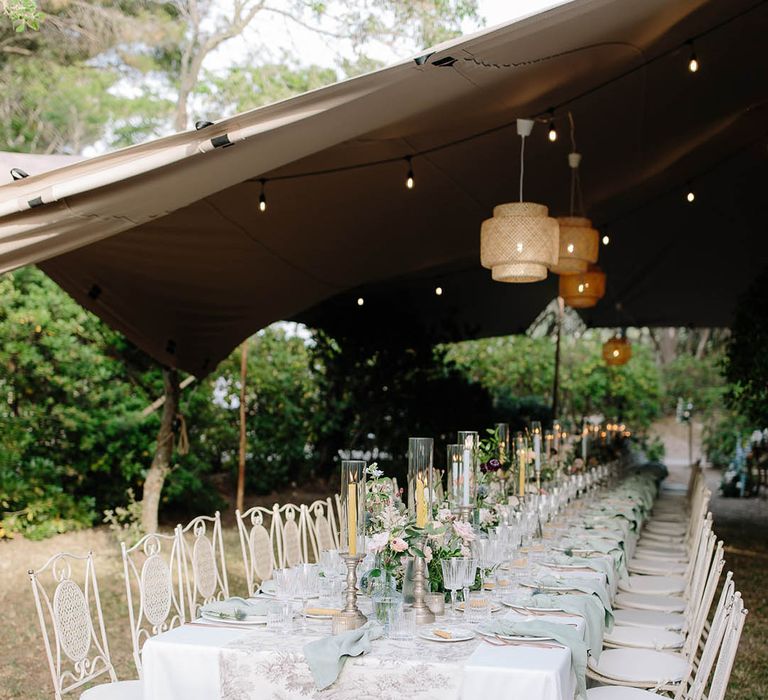 Beautiful Italian tablescape with ribbed glassware, gold candlestick holders and flower arrangements that include thistles, peonies, carnations and wildflowers under a marquee with rattan lamps