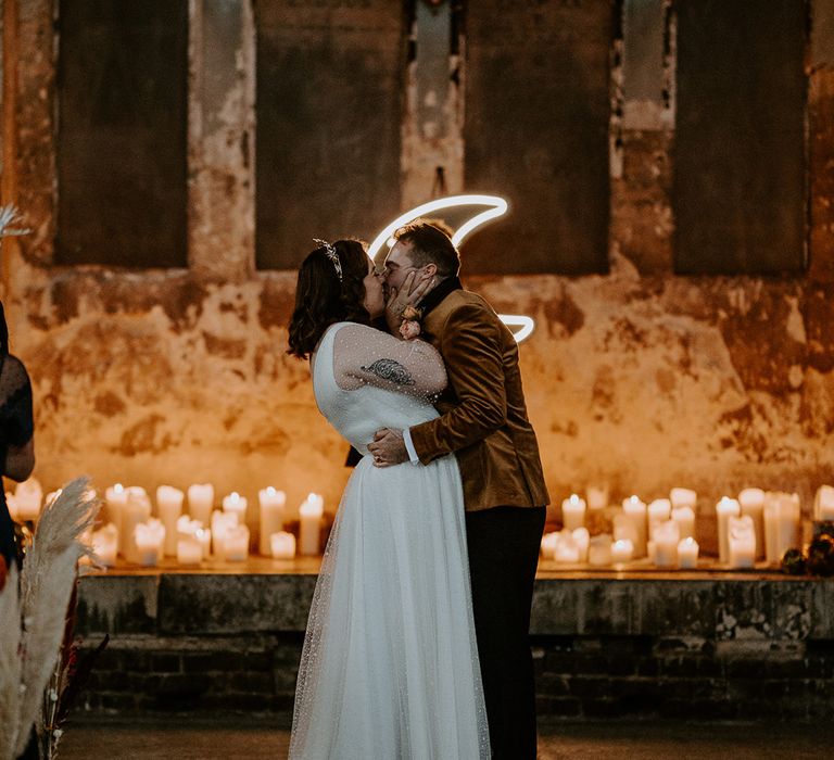 Bride and Groom share a kiss at unique wedding venue. Their wedding lighting is created by candles and a moon neon sign