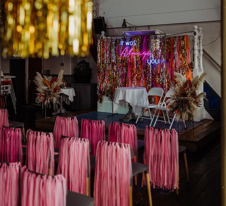 Pink, orange, gold and silver metallic streamer backdrop with pink streamers on the back of chairs, gold streamers on the lights and fairly lights hung up on the ceiling  